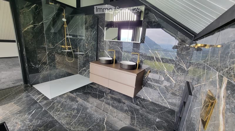 Modern bathroom with dark marble tiles, double sink vanity, gold fixtures, and a glass-enclosed shower area.