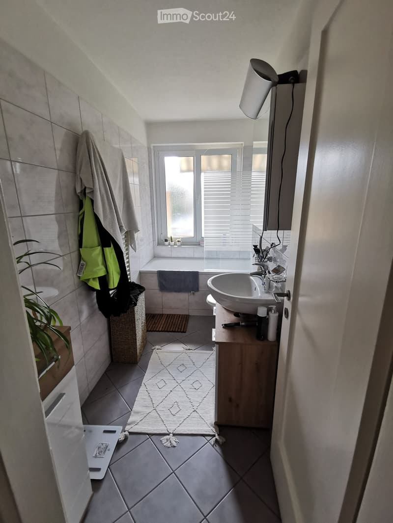 Modern bathroom with tiled walls and floor, featuring a sink with a faucet, a bathtub, a mirror, a towel hanging on the wall, a small plant, a wicker basket, and a patterned rug.