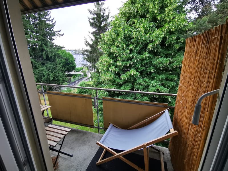 Balcony with a wooden chair, small table, bamboo screen, and metal railing overlooking a green area with trees.