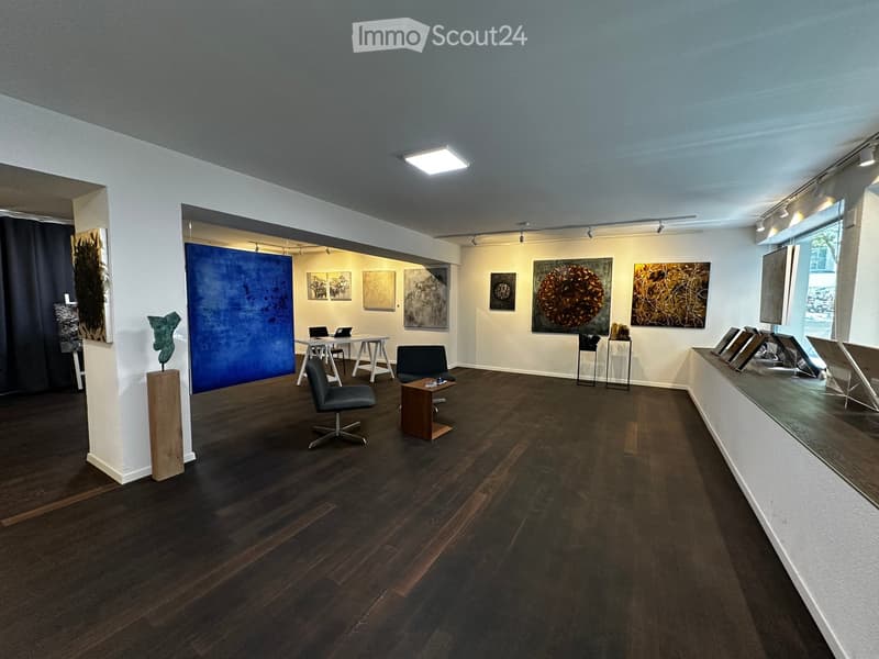 Modern gallery space with dark wooden flooring, white walls, and large artworks displayed. Includes a small table with laptops, chairs, and a sculpture on a pedestal.