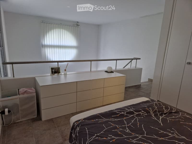 Modern bedroom with a bed, nightstand, drawer unit, and a small picture frame on the nightstand. The room features a balcony railing and a window with blinds.