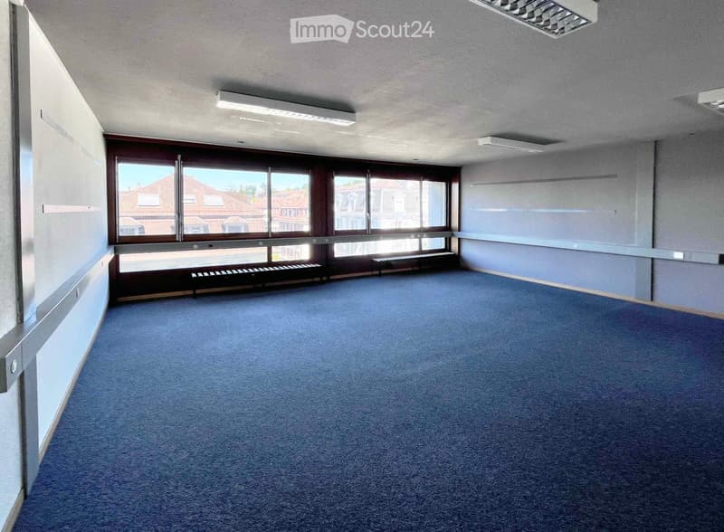 Empty room with blue carpet, large windows, and minimal furnishings including a small bench and a low cabinet.