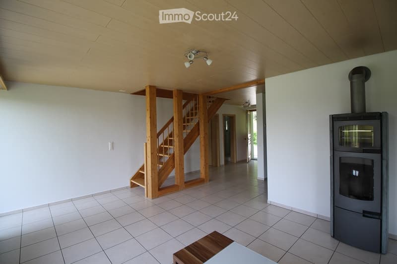 The image shows a modern, well-lit living space with a wooden staircase leading to an upper level, a wooden bench, and a wood-burning stove with a chimney. The room features white walls, a wooden ceiling, and tiled flooring.
