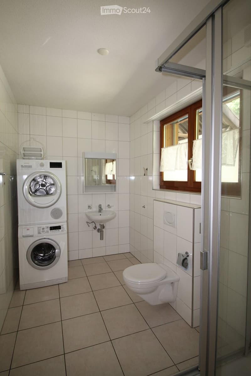 Modern bathroom with white tiles, a washing machine, toilet, sink, and mirror. Features a window with wooden frame and white curtains.