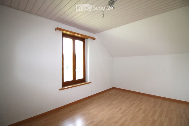 Empty room with wooden flooring, white walls, and a large window with wooden frame and built-in shelf. The ceiling has exposed wooden beams and a modern light fixture.