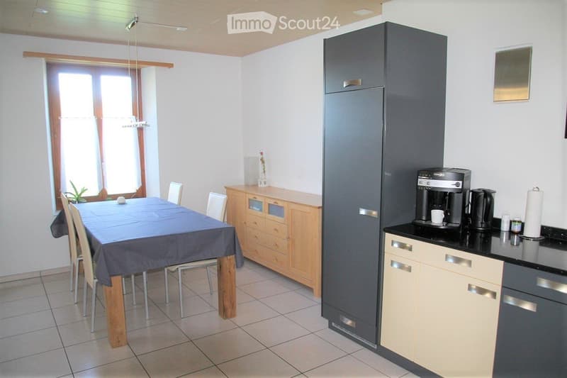 A modern kitchen and dining area with a dining table, chairs, a refrigerator, coffee maker, and a wooden cabinet.