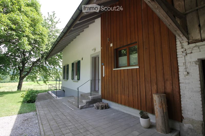 A two-story house with a mix of wooden and plaster exterior walls, featuring a covered entrance with steps, a small garden area, and a view of a green field and trees.