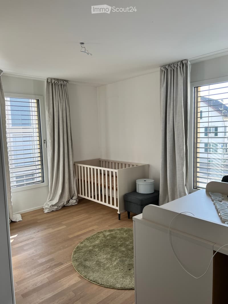 A well-lit nursery with a wooden crib, a small grey stool, a round green rug, and a white table with baby items.