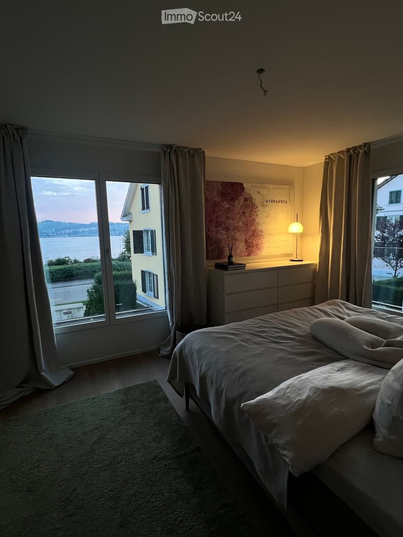 Modern bedroom with a large bed, nightstand, lamp, and scenic view through large windows.
