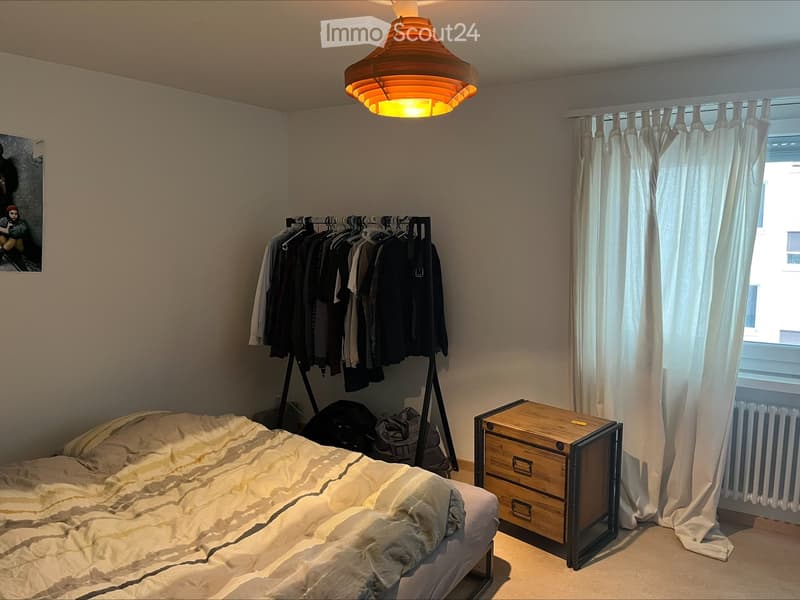 Cozy bedroom with a bed, nightstand, clothing rack, and a picture frame on the wall. The room features a large window with white curtains and a modern hanging lamp.