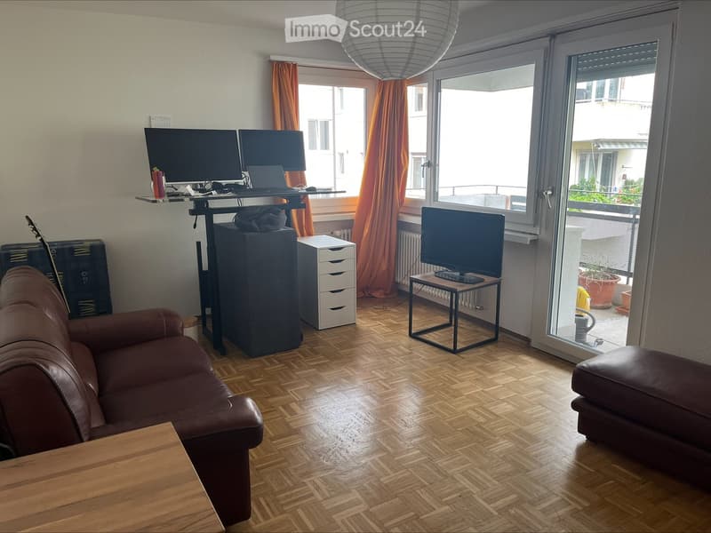 A modern living room with a brown leather sofa, a wooden coffee table, a small black table with a TV, a laptop on a standing desk, and a guitar leaning against the wall. The room has wooden flooring and large windows with orange curtains, leading to a bal