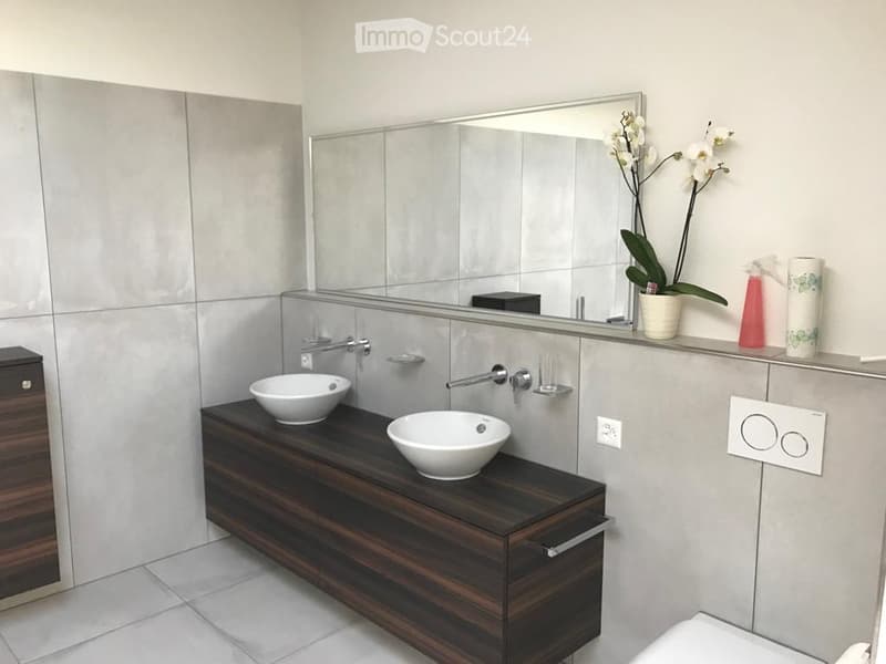 Modern bathroom with a dark wooden vanity, two white sinks, a large mirror, and decorative elements including a flower vase and soap dispenser.