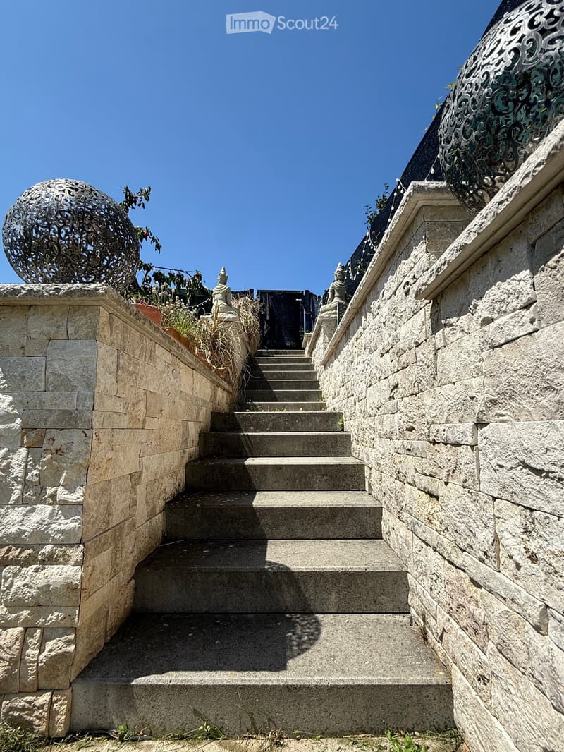 Stone staircase leading up with decorative metal sphere and small statues on either side, surrounded by stone walls and greenery.