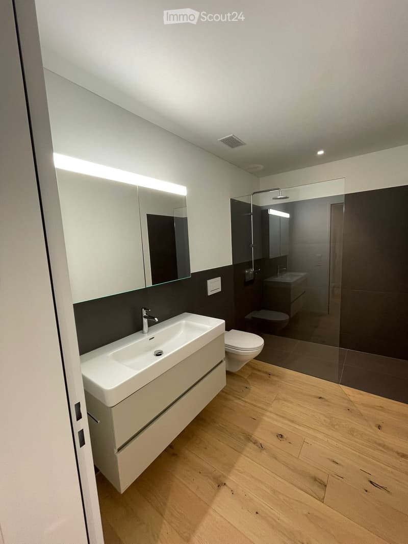 Modern bathroom with a white sink, chrome faucet, toilet, large mirror, and glass-enclosed shower area. Light wood flooring and sleek design.