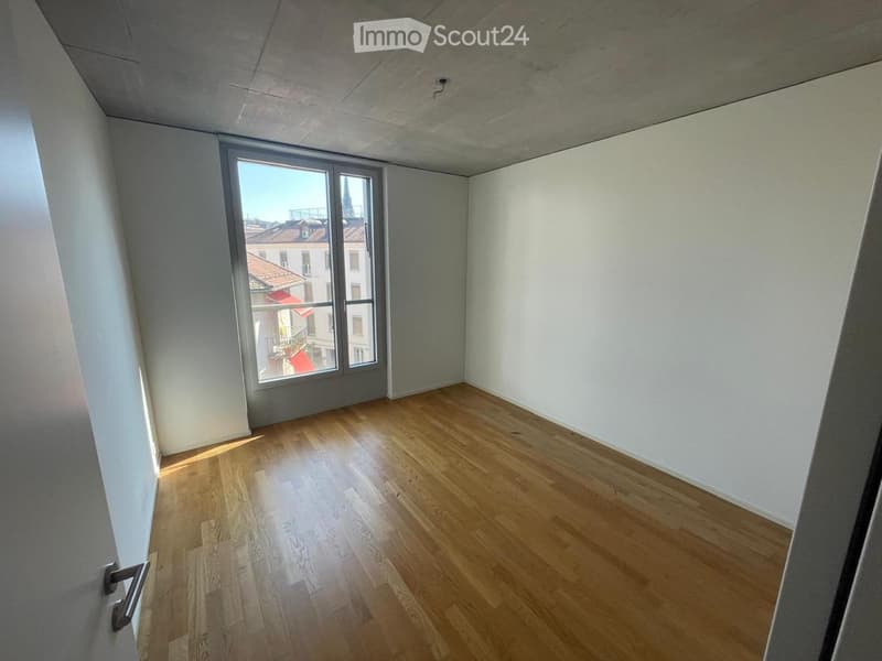 Empty room with wooden flooring, white walls, and a large window offering a cityscape view.