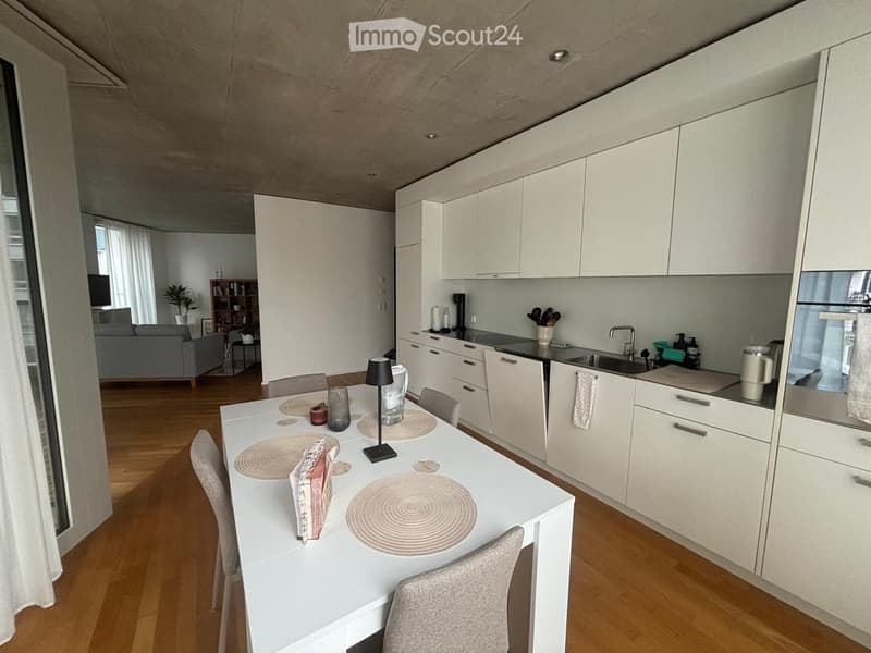 Open-plan living and dining area with modern white kitchen cabinets, a dining table with chairs, a lamp, and a sofa in the background.