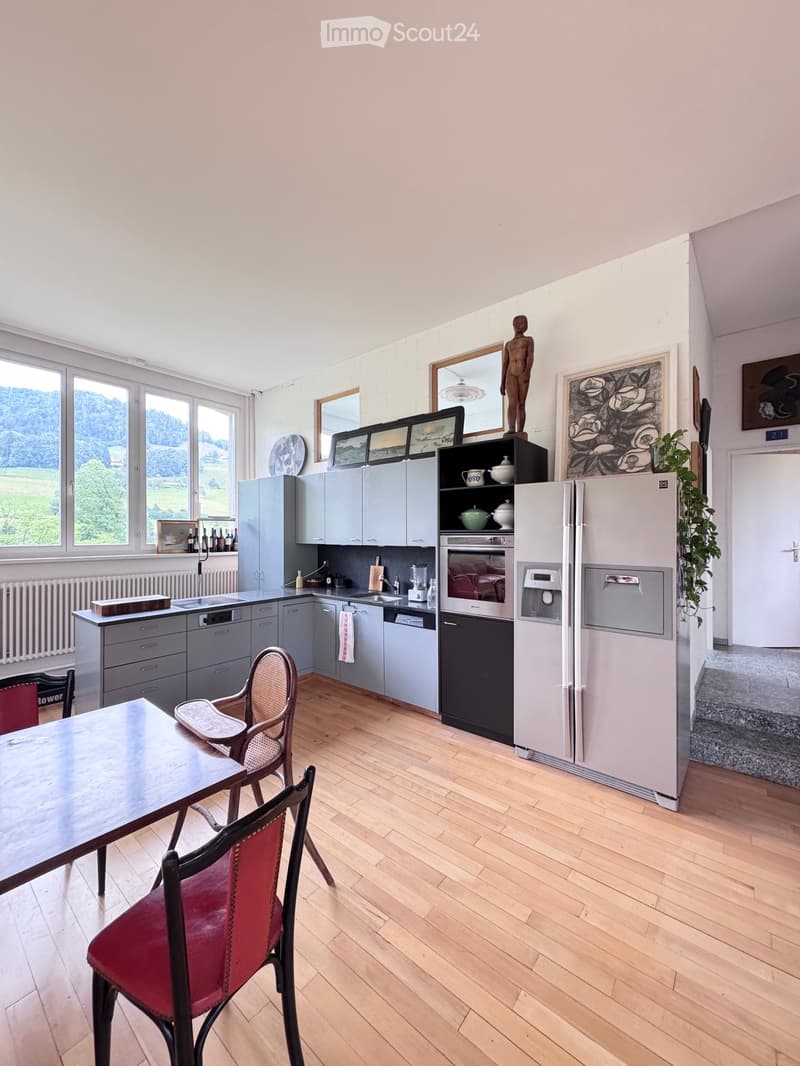 Modern kitchen with grey cabinets, stainless steel appliances, wooden floor, large windows with scenic view, dining table with chairs, and decorative items on shelves.