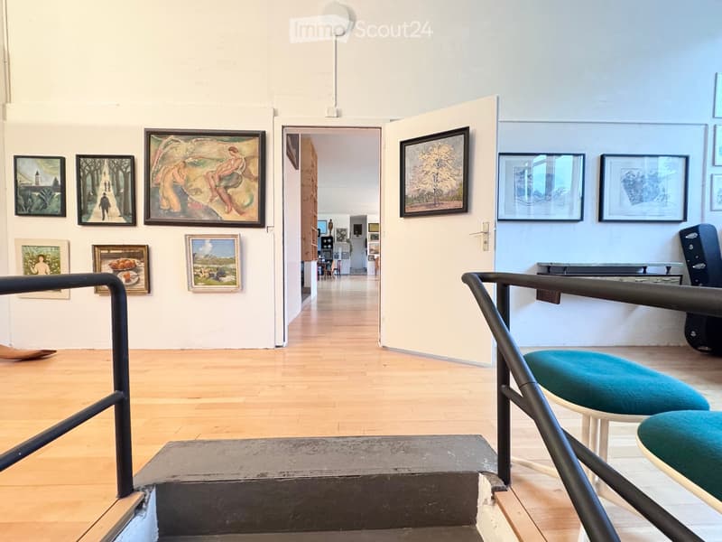 Gallery space with wooden floors, white walls, and several paintings displayed. Two teal stools are placed near a staircase with a metal railing.