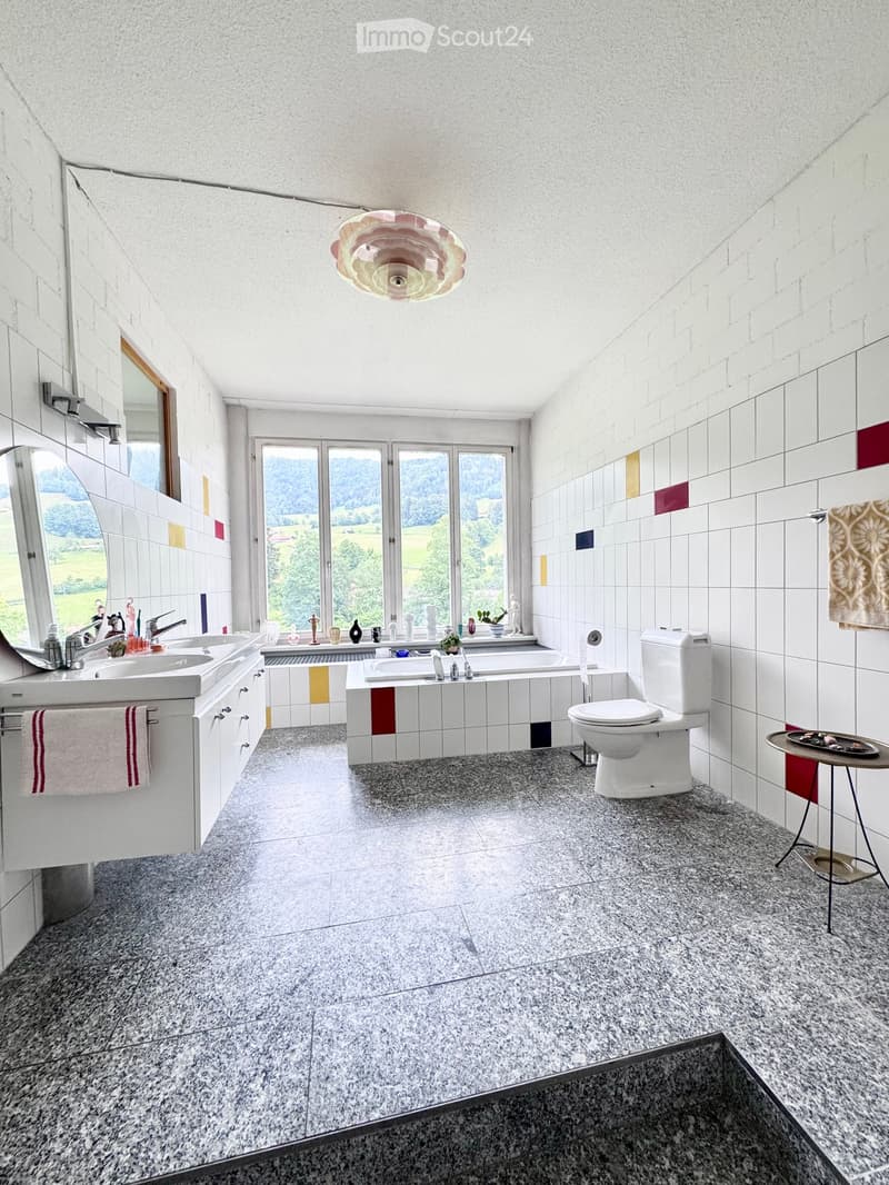 Modern bathroom with white tiles, a large window offering a scenic view, a double sink, a bathtub, a toilet, and decorative items on the windowsill.