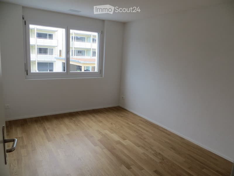 Empty room with light wooden flooring and white walls, featuring a large window with white frames and a view of neighboring buildings.