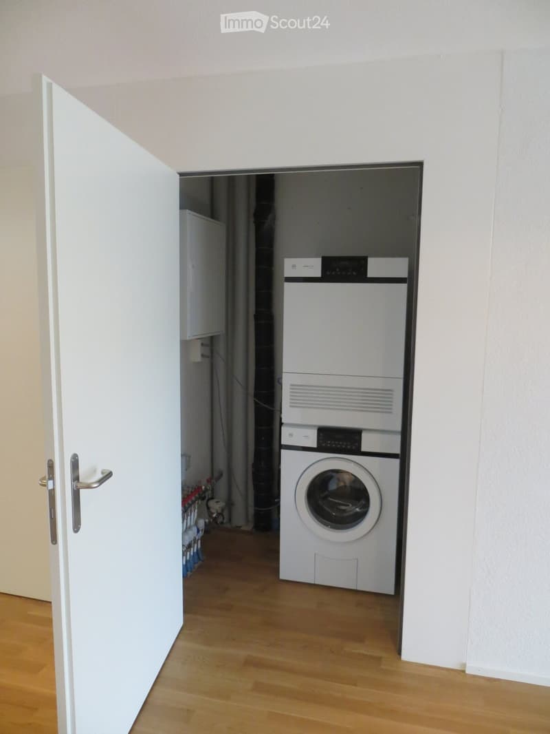A small, well-lit room with a wooden floor, containing a washing machine and a dishwasher stacked vertically.