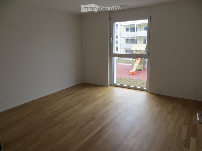 Empty room with wooden flooring and large window opening to a playground area outside.