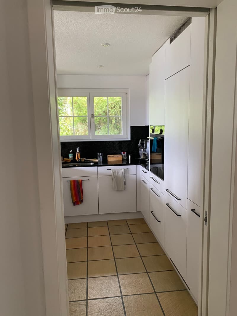 Modern kitchen with white cabinets, tiled floor, and a window overlooking greenery. Includes a sink, oven, coffee maker, and towels.