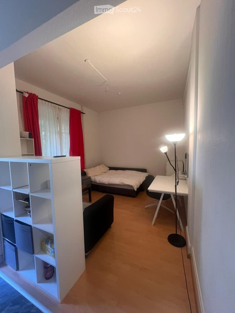 A small, well-lit bedroom with a bed, a chair, a desk, a shelf, and a lamp. The room features wooden flooring and white walls.