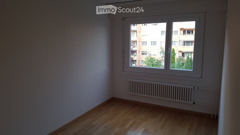 Empty room with wooden flooring, white walls, a window with white frame, and a radiator below the window.
