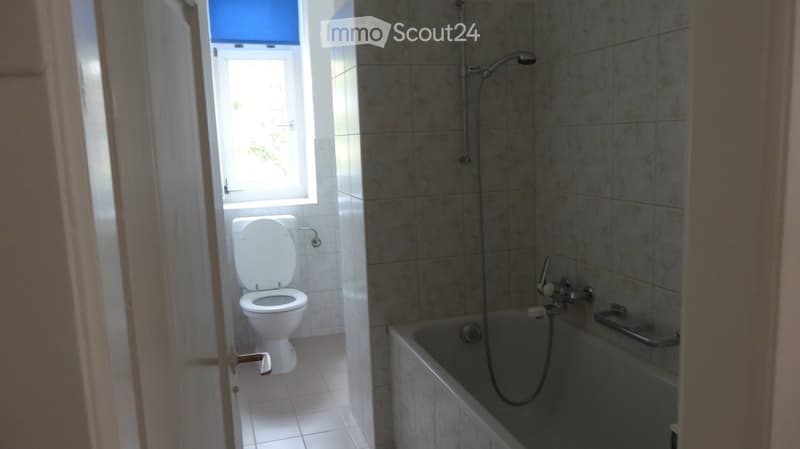 Bathroom with a toilet, bathtub, and showerhead. The room has tiled walls and a window letting in natural light.