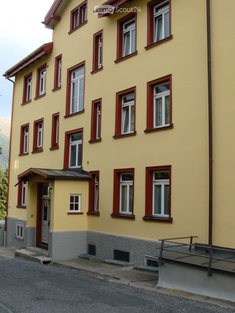 Two-story yellow house with red window frames and a gray stone base. Features multiple windows, a small porch with steps, and a metal railing on the right side.