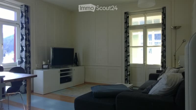 Living room with a grey sofa, wooden table, chairs, television on a white cabinet, and a floor lamp near the window.