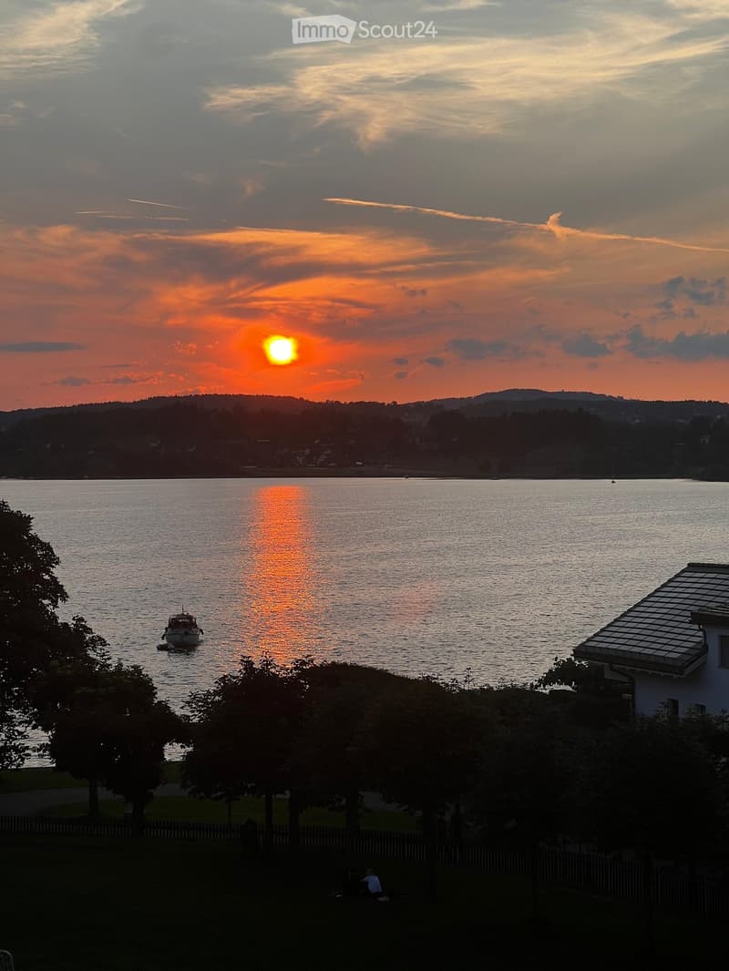 View of a lake at sunset with a boat on the water, distant hills, and a house partially visible on the right side.