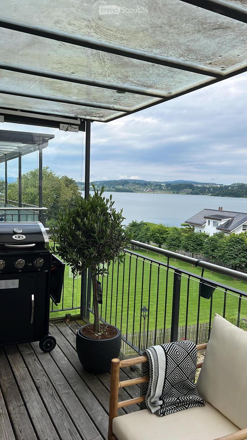 Balcony with wooden decking, a modern grill, a potted plant, and a comfortable chair with a throw blanket, overlooking a scenic lake view.