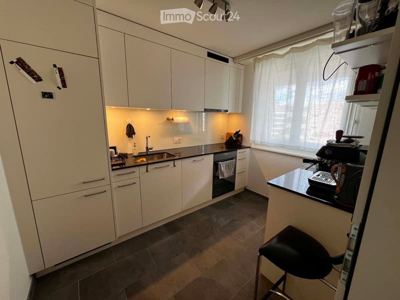 Modern kitchen with white cabinets, black countertops, stainless steel sink, coffee maker, and a black stool.