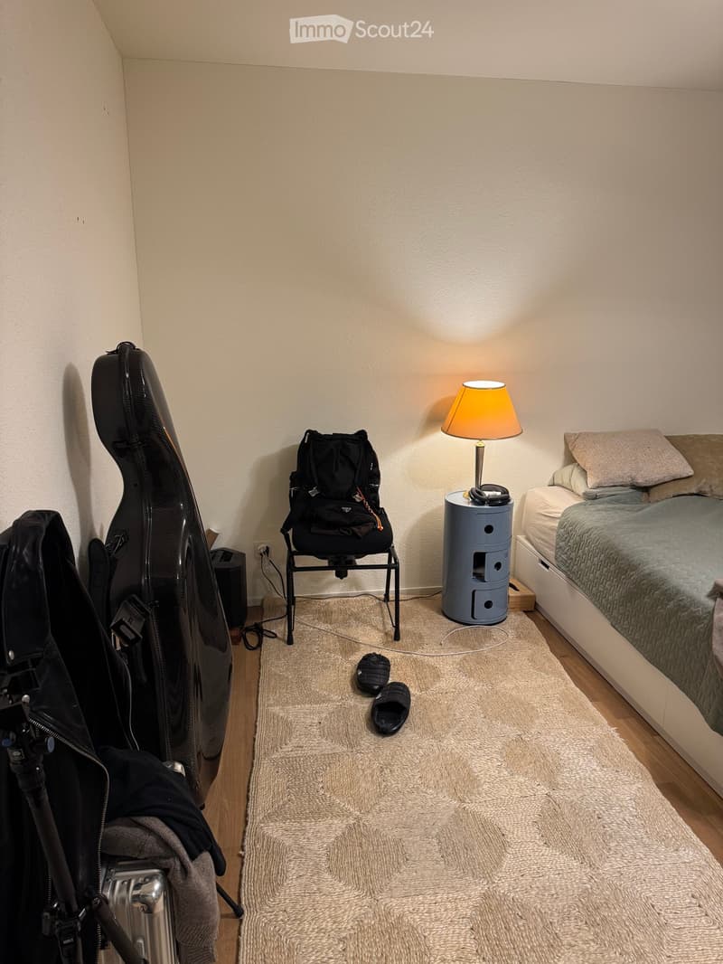 Cozy bedroom with a bed, a small lamp on a side table, a chair with a backpack, a guitar case, and a camera tripod near the door.