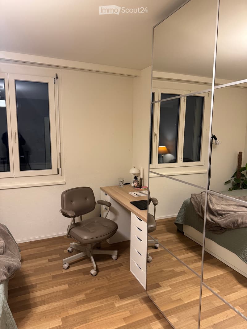 Modern bedroom with a large mirror, a desk with a chair, a laptop, a lamp, and a cup. The room features wooden flooring and large windows.