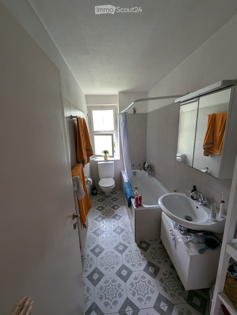 Bathroom with a toilet, bathtub, sink, mirror, and towels. Features a window with a small plant on the windowsill and a patterned tile floor.