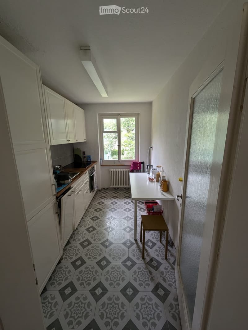 The kitchen features white cabinets, a dark countertop, a sink, an oven, a table with a stool, and a window with a view of greenery outside. The floor has a patterned tile design.