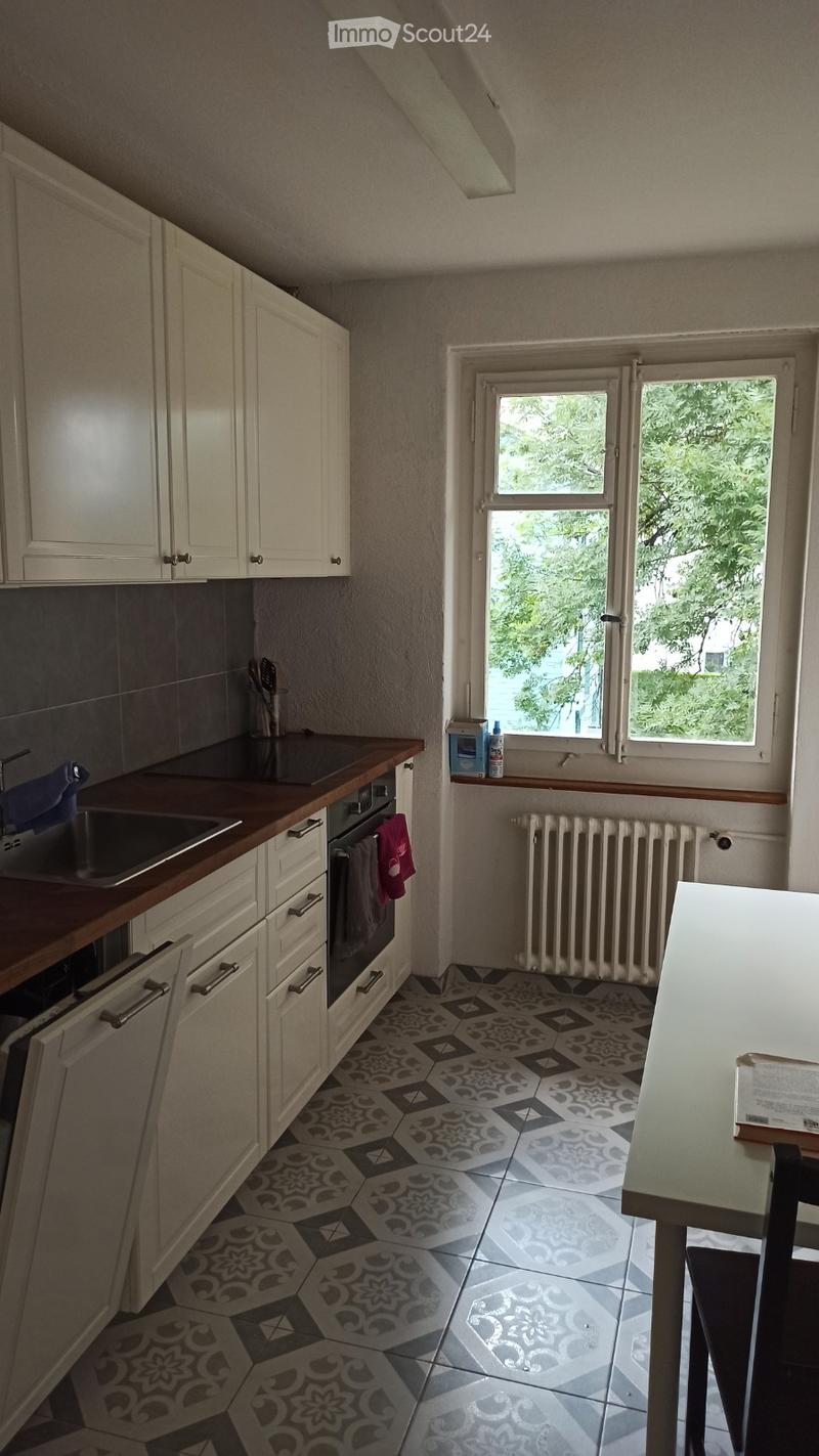 Bright kitchen with white cabinets, dark countertops, and a patterned tile floor. Features a sink, oven, dishwasher, and a small table near the window.