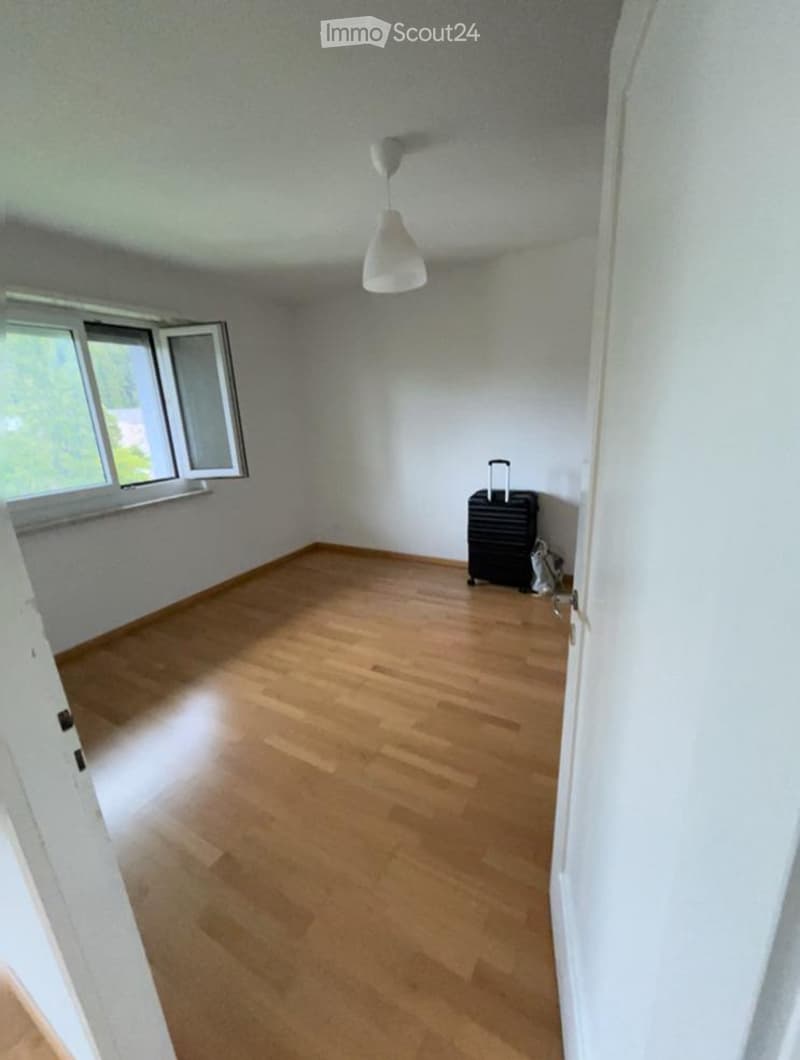 Empty room with wooden flooring, white walls, and a single window with white frame. A ceiling light fixture is visible, and a small black suitcase is placed near the window.