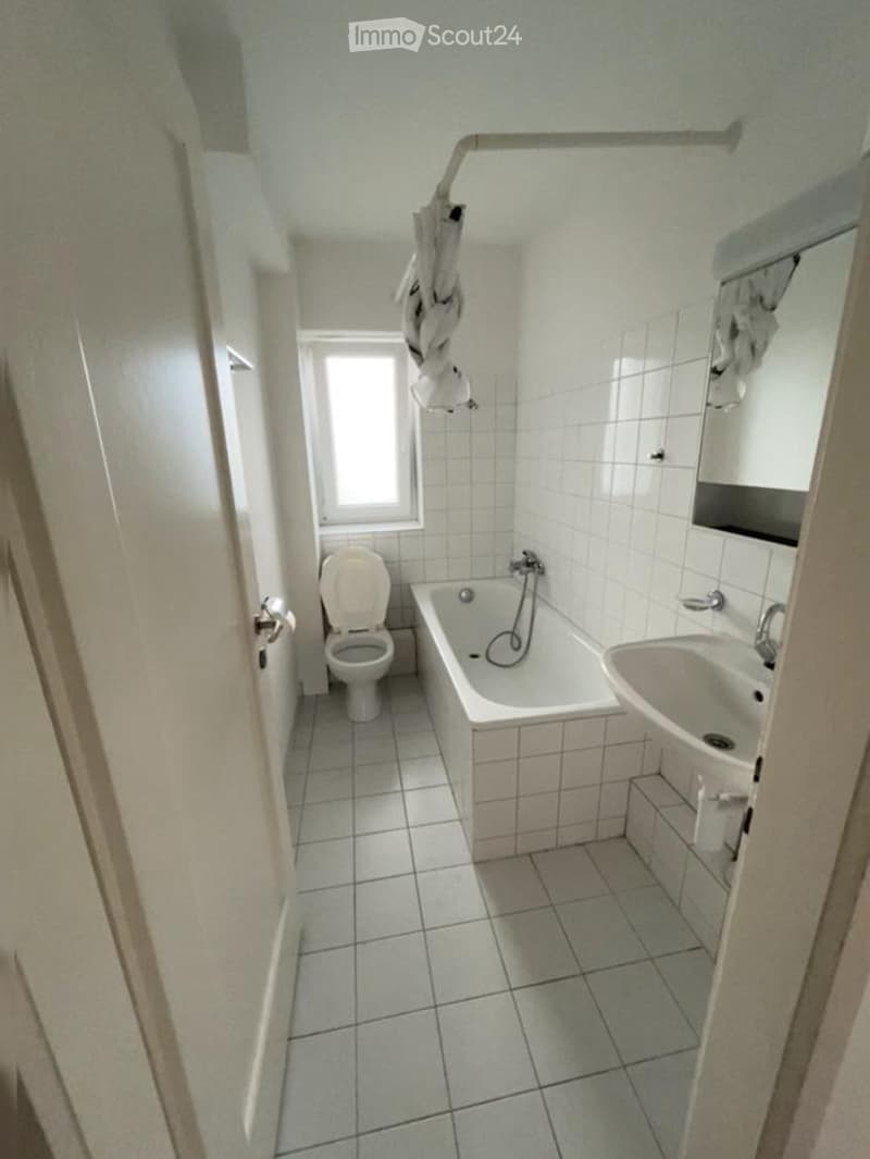 A small bathroom with white tiled walls and floor, featuring a toilet, bathtub, sink, and a mirror above the sink. A window provides natural light, and a towel hangs on the wall.