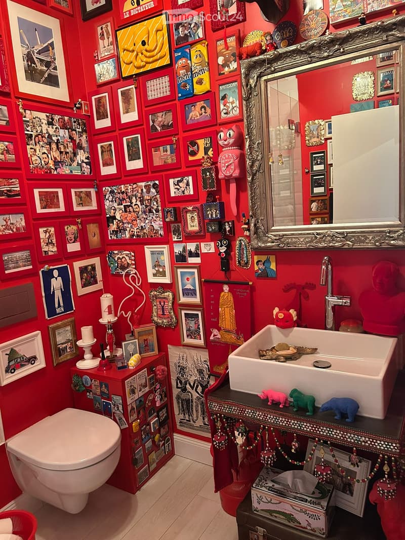 A vibrant red bathroom with a toilet, sink, and ornate mirror. The walls are covered with numerous framed pictures and posters, creating a colorful and eclectic atmosphere. A small cabinet holds various decorative items, including a clock, candle, and sma