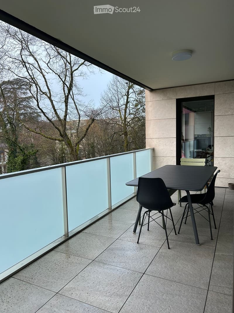 Modern balcony with a glass railing, tiled floor, and a small black table with two chairs.