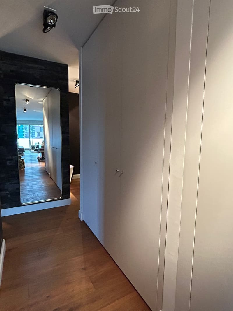 A narrow hallway with wooden flooring, white walls, and a large mirror reflecting a view of another room with furniture and a window.