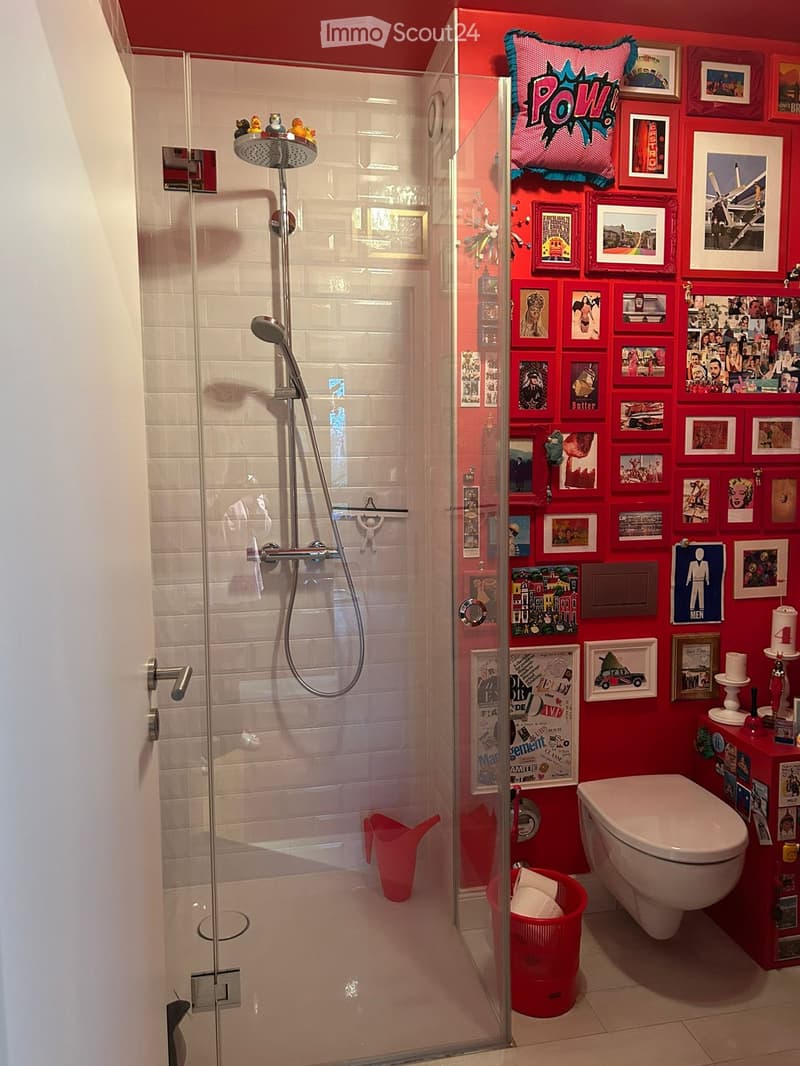 Modern bathroom with glass shower enclosure, white tiles, red walls, decorative pictures, white toilet, red storage box, red bucket, and shower head.