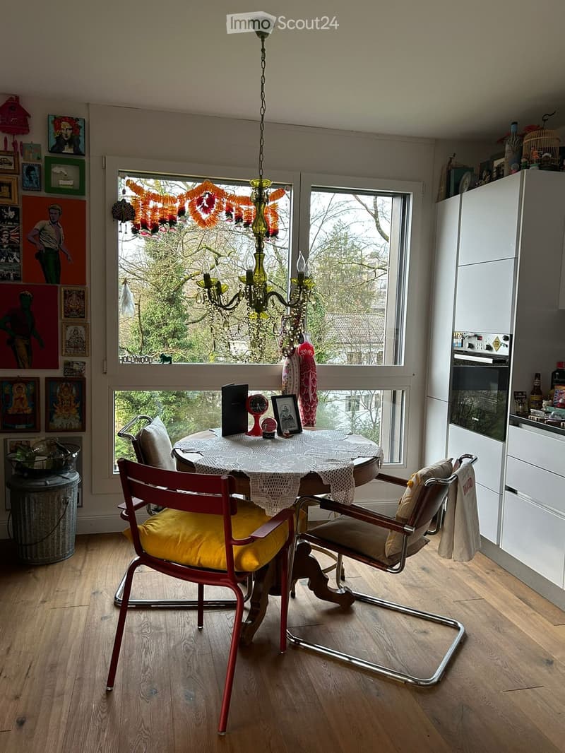 Dining room with a round table, chairs, a chandelier, and a window with a view of trees outside.