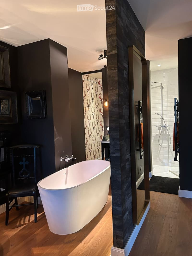 Modern bathroom with a freestanding bathtub, dark walls, wooden flooring, a glass-enclosed shower, and a small table with a mirror.