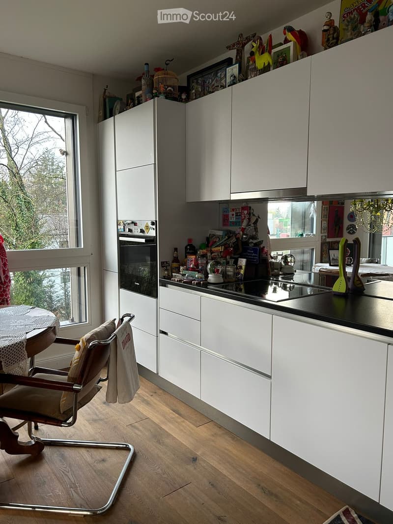 Modern kitchen with white cabinets, wooden floor, and a large window. Features a chair, table, oven, fridge, and various decorative items on the counter and shelves.