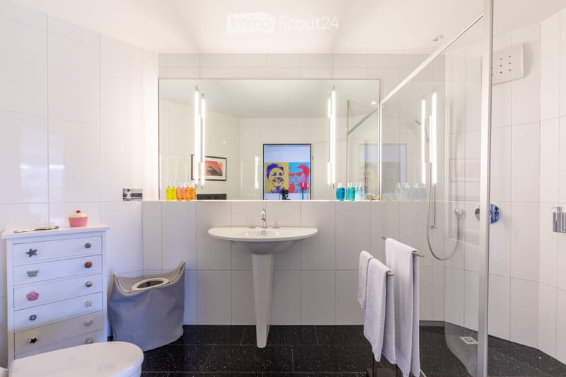 Modern bathroom with white tiles, a large mirror, a sink, a toilet, a shower, and a towel rack with towels. There is a framed picture on the wall and a small cabinet with a cake on top.